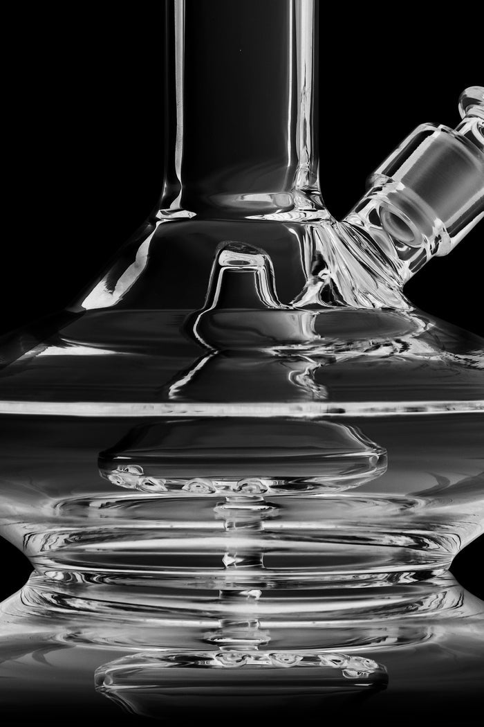 Clear glass water pipe (bong) with intricate design, reflecting in a dark surface.
