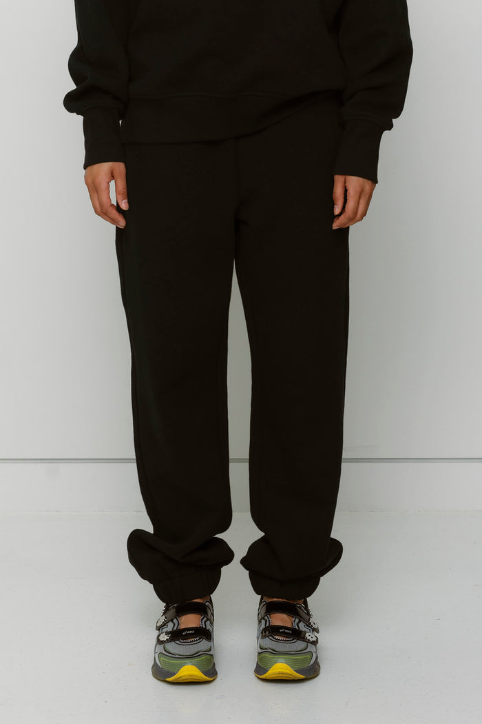 Model displays black sweatpants and matching sweatshirt with Asics sneakers against a white wall.