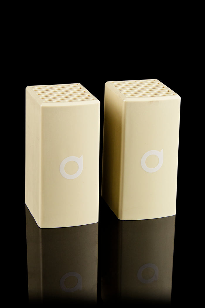 Two cream-colored cannabis vaporizers with a honeycomb top and circular logo, displayed on a reflective surface."